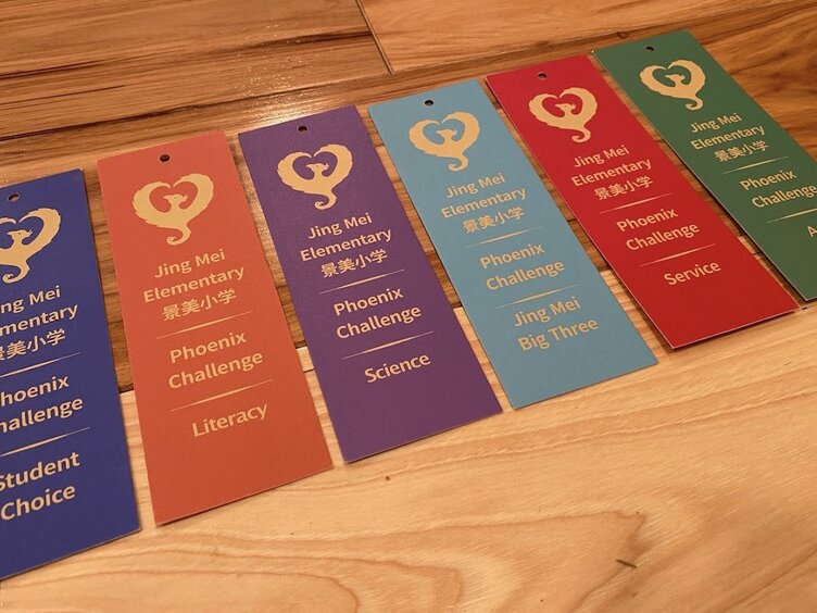 Six Phoenix Challenge bookmarks fanned out on a wooden table, one per category — Student Choice, Literacy, Science, Jing Mei Big Three, Service and Art — each printed with the Jing Mei Elementary 景美小学 phoenix mark.