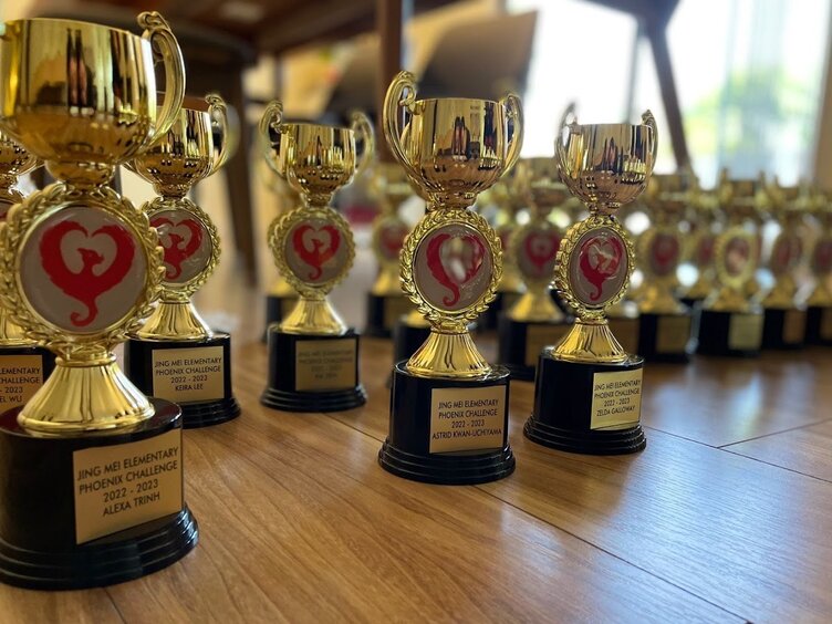 A row of gold Phoenix Challenge trophies on a wooden table, each engraved with a student's name and topped with the red Jing Mei phoenix emblem.